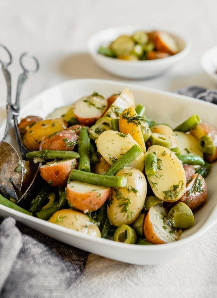 Side angle image of green beans, potatoes, lives and herbs in a white square dish set on a table.