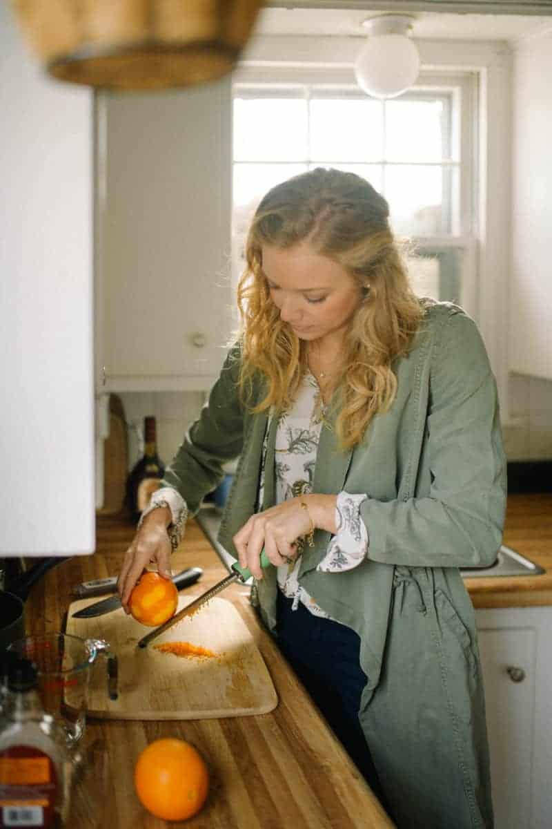 photograph of woman in a green coat in a kitchen zesting an orange
