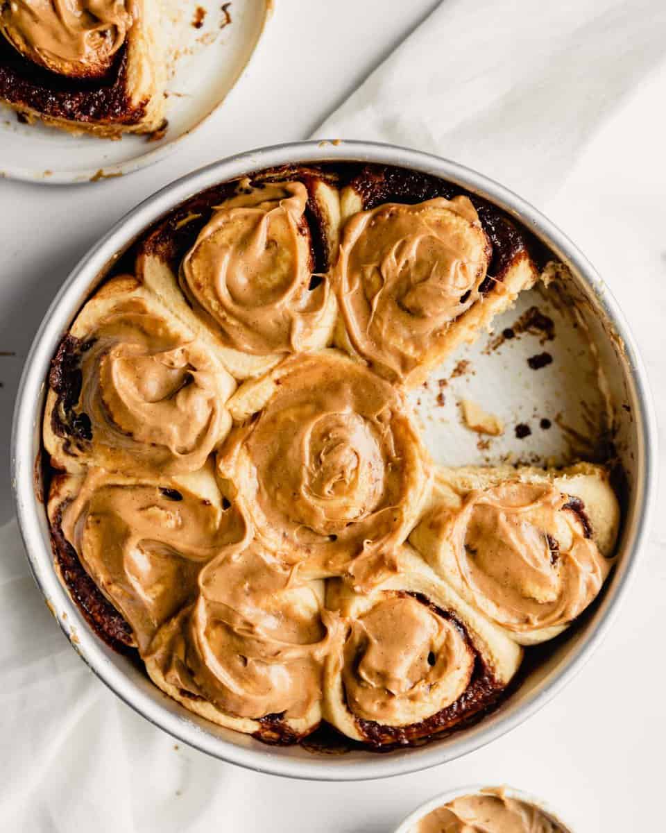 overhead image of baked and frosted cinnamon rolls in a round cake pan on a white wooden table with extra frosting set off to the side