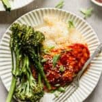 chicken breast coated in a red sauce plated over rice with broccolini in an off-white bowl