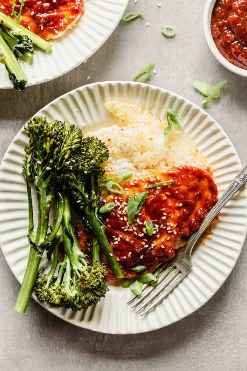 chicken breast coated in a red sauce plated over rice with broccolini in an off-white bowl