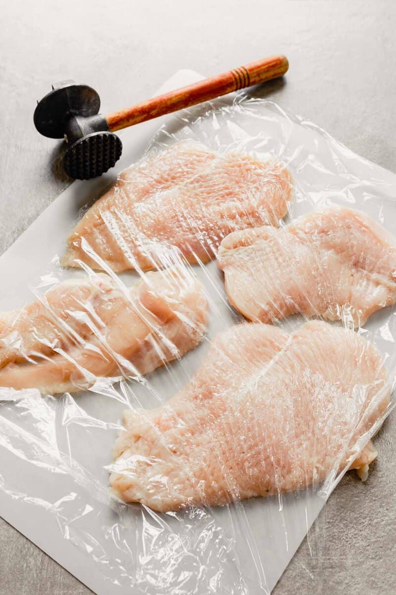 raw chicken breast on cutting board covered with plastic wrap