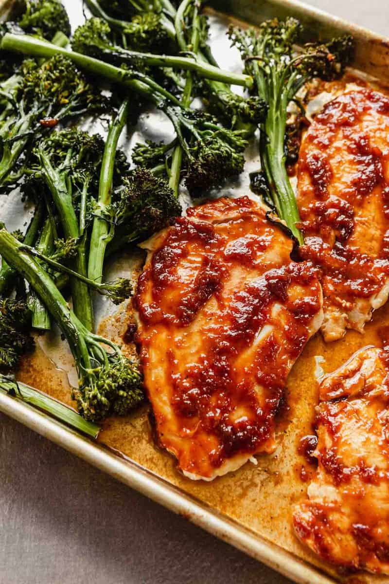 cooked chicken on a baking sheet coated in a red sauce with broccolini arranged around