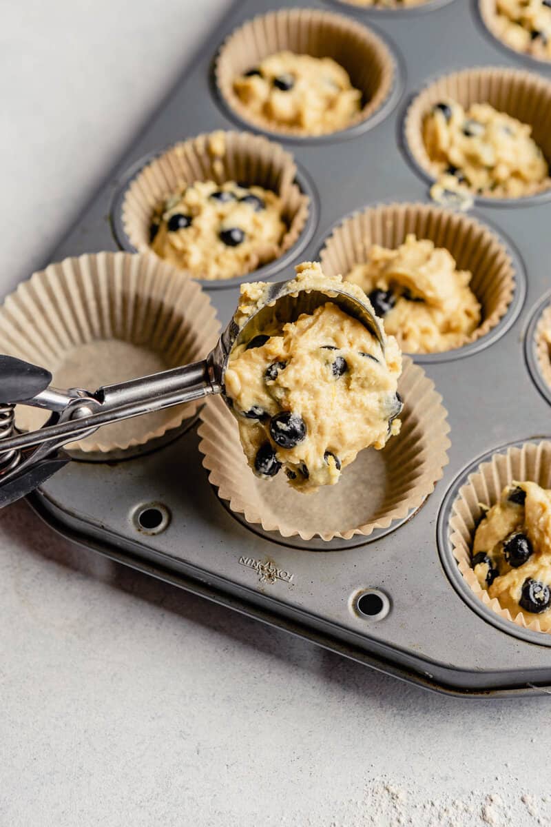Healthy blueberry muffin batter getting scooped into paper-lined muffin tin.