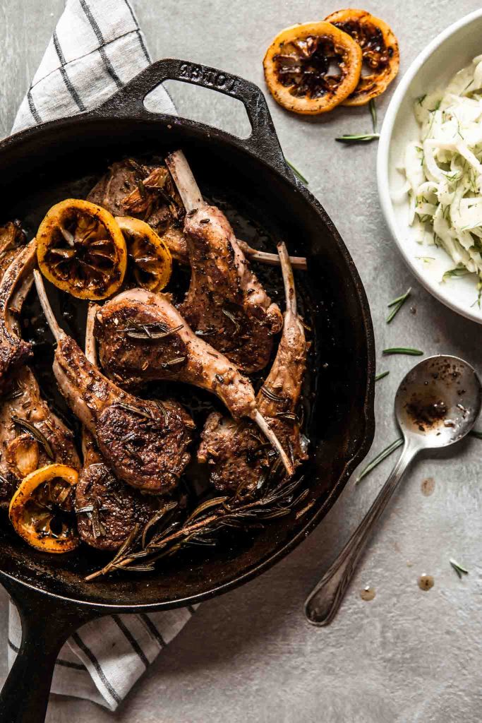 Overhead image of lamb chops in a cast-iron skillet set on top of a stripped towel with a spoon set to the bottom corner and a fennel salad set off to the top right corner