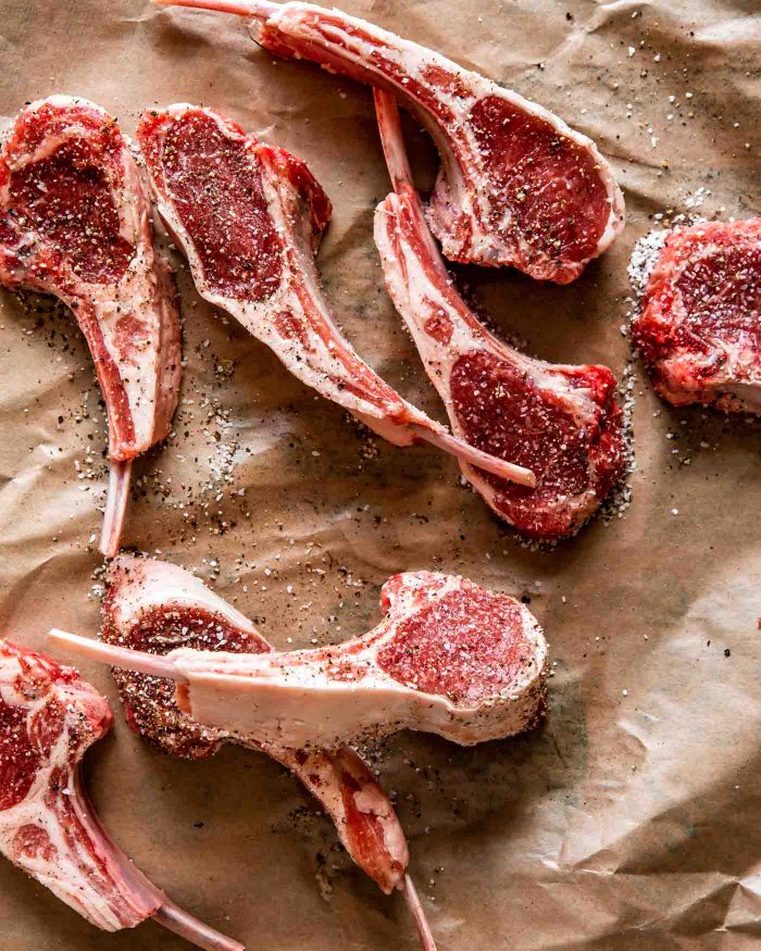Overhead image of raw lamb rib chops set in a brown piece of butcher paper; seasoned with salt and pepper