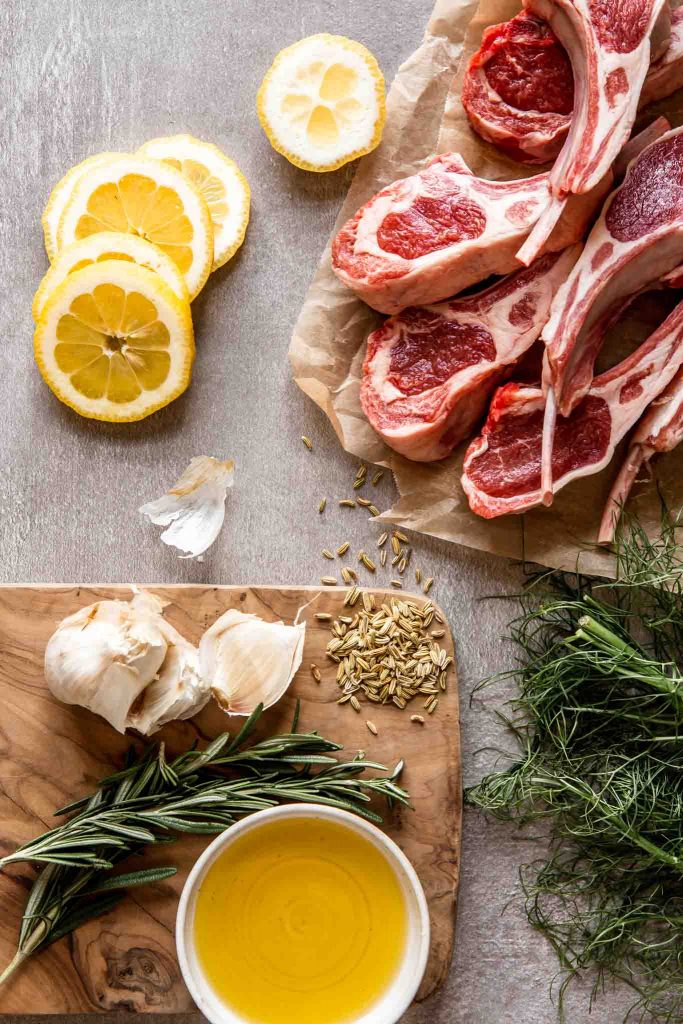 overhead image of raw lamb rib chops, lemon slices, garlic, fennel seeds, fresh rosemary and olive oil arranged on a counter top