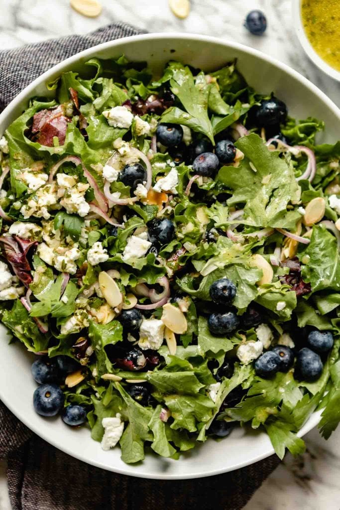 image of a large white bowl filled with salad greens, blueberries, shallot, almonds, and goat cheese