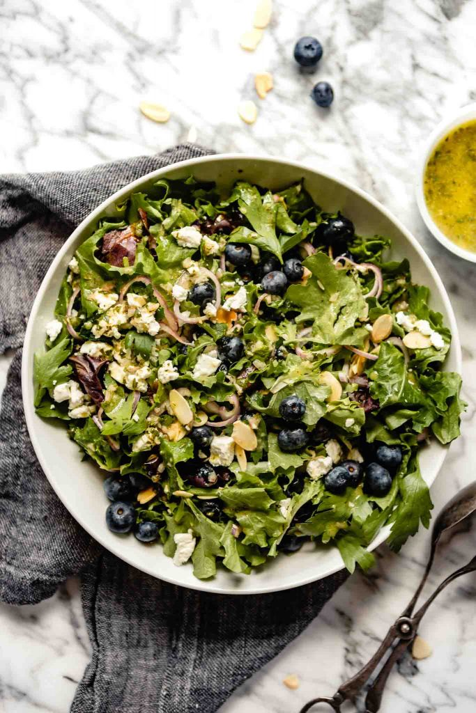 image of a large white bowl filled with salad greens, blueberries, shallot, almonds, and goat cheese