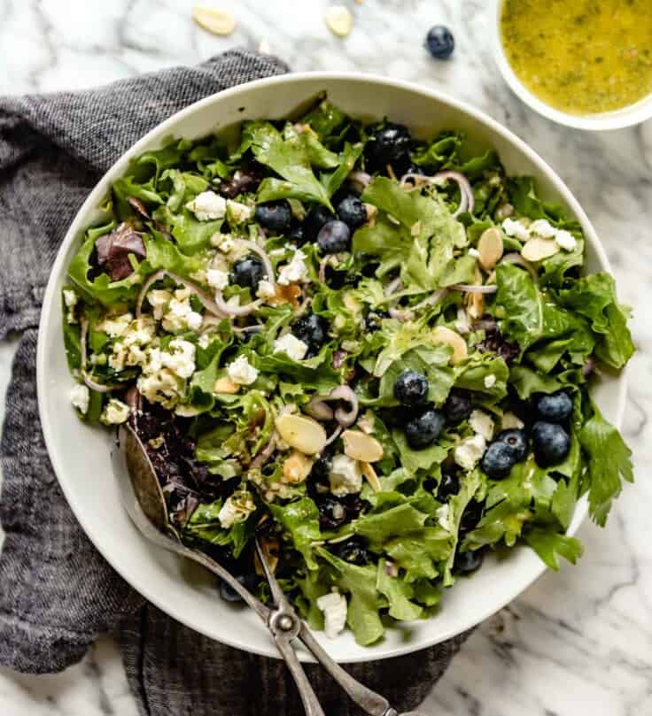 image of a green salad in a large white bowl topped with blueberries, almonds, cheese and sliced shallots