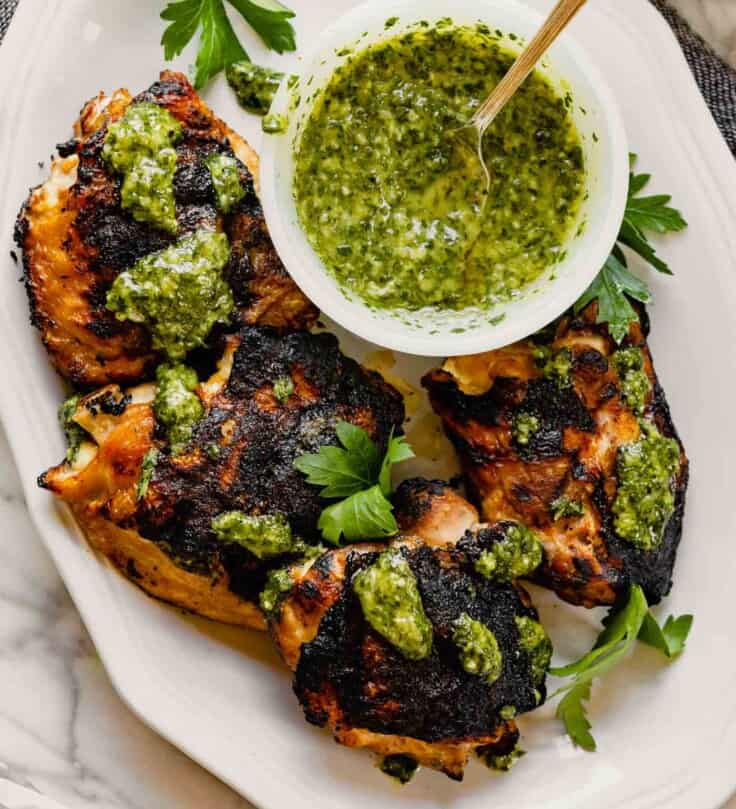 Grilled chicken thighs on a white plate drizzled with chimichurri sauce with a bowl of green sauce off to the side