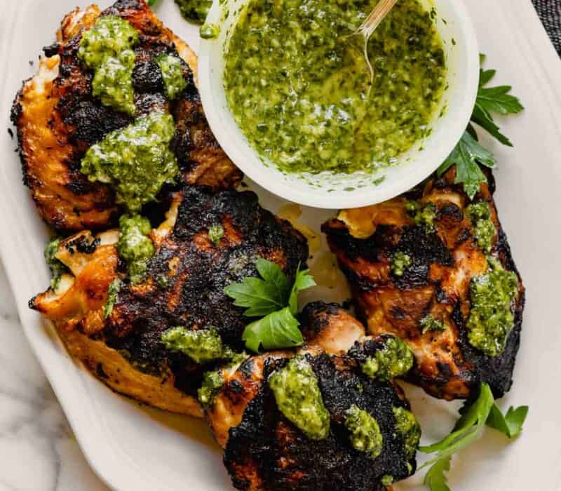 Grilled chicken thighs on a white plate drizzled with chimichurri sauce with a bowl of green sauce off to the side