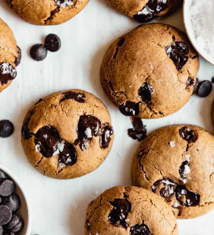chocolate chip cookies arranged on parchment paper with a bowl of chocolate chips to the bottom left and salt to the top right