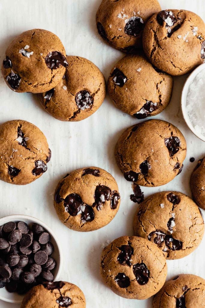chocolate chip cookies arranged on parchment paper with a bowl of chocolate chips to the bottom left and salt to the top right