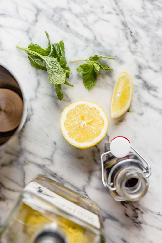 image of a cocktail shaker, bottle of whiskey, maple syrup, lemon wedges and fresh mint arranged on a marble table.