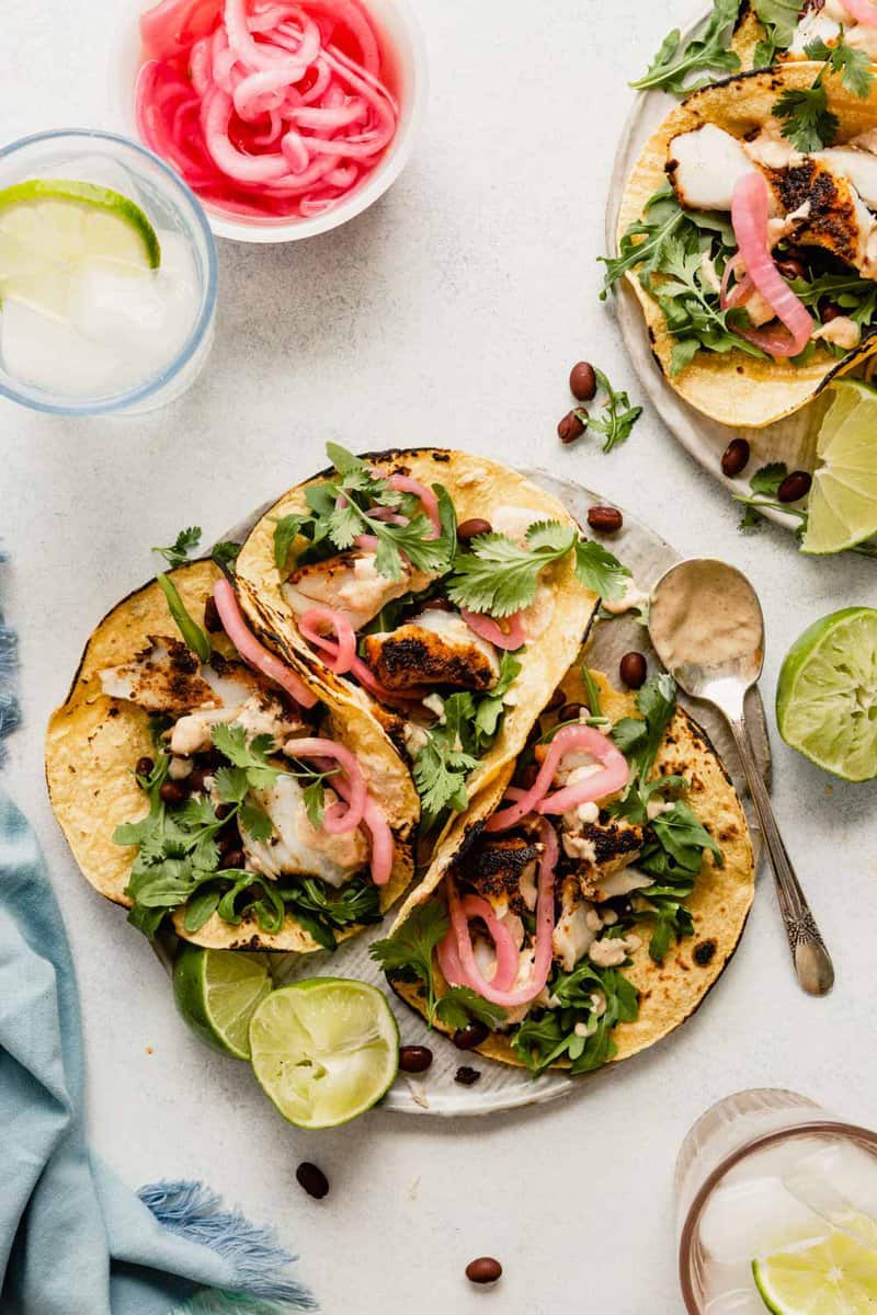 three corn tortillas filled with blackened fish, arugula, black beans and a creamy sauce on a plate with picked onions and bubbly water set around