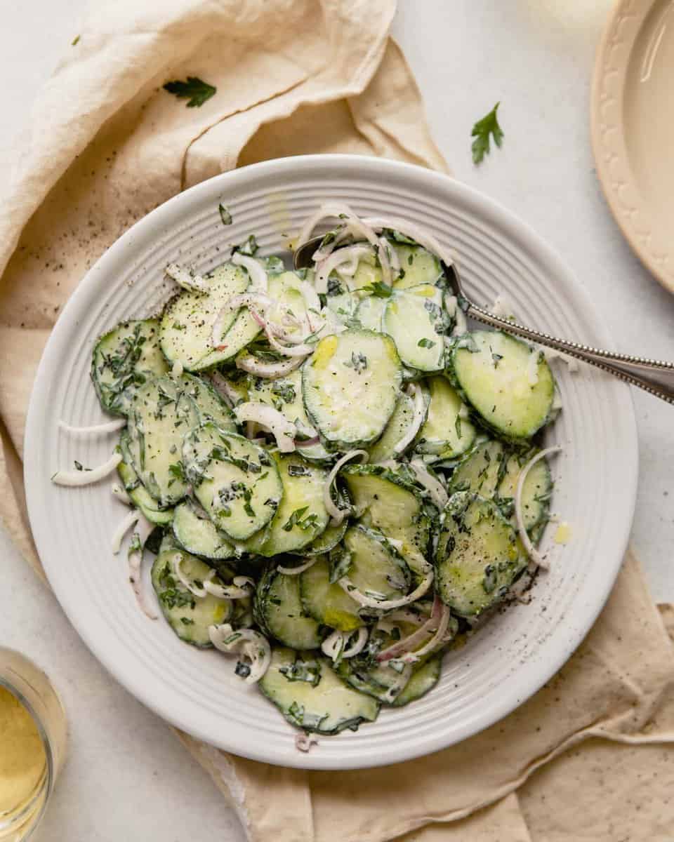 thinly sliced cucumber dressed in a creamy dressing in a white bowl set on top of a yellow napkin