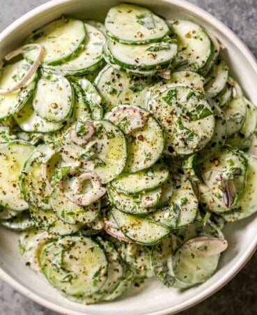 Thinly sliced cucumbers and shallot coated in a creamy Greek yogurt dressing with chopped herbs in a shallow white bowl.