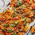 nachos on a baking sheet topped with cilantro, tomatoes, kimchi, melted cheddar cheese and a sauce