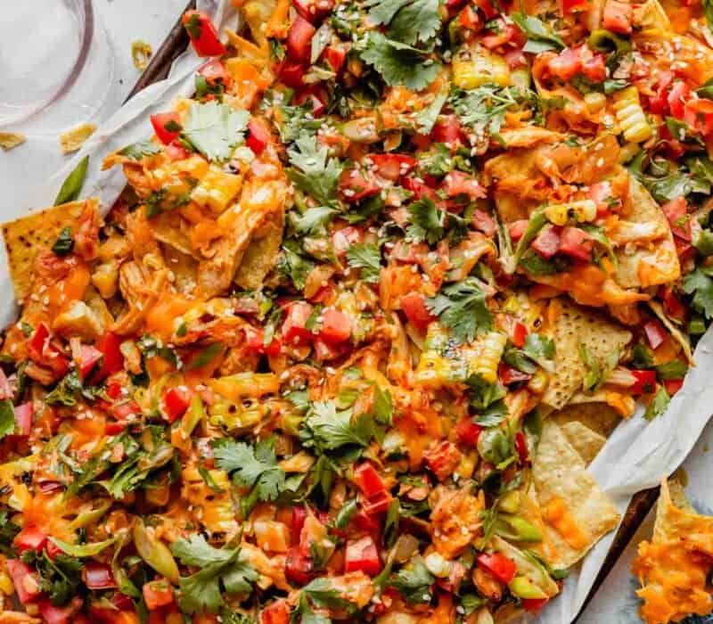nachos on a baking sheet topped with cilantro, tomatoes, kimchi, melted cheddar cheese and a sauce