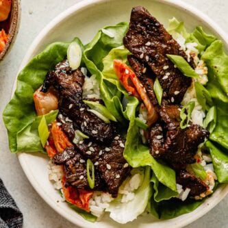 Strips of sliced beef bulgogi in lettuce wraps with rice, kimchi and sesame seeds. Set in a shallow white bowl.