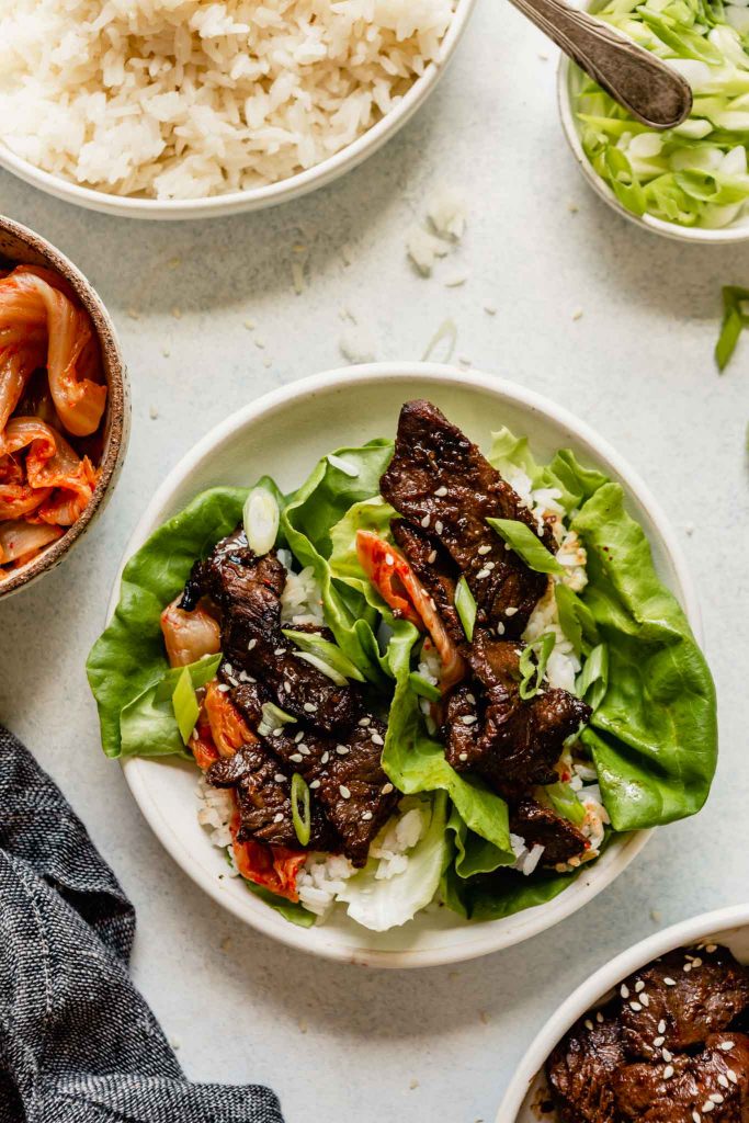 white bowl with lettuce wraps filled with rice, kimchi and grilled beef bulgogi. Set on a table with bowls of rice, scallions, and kimchi set around