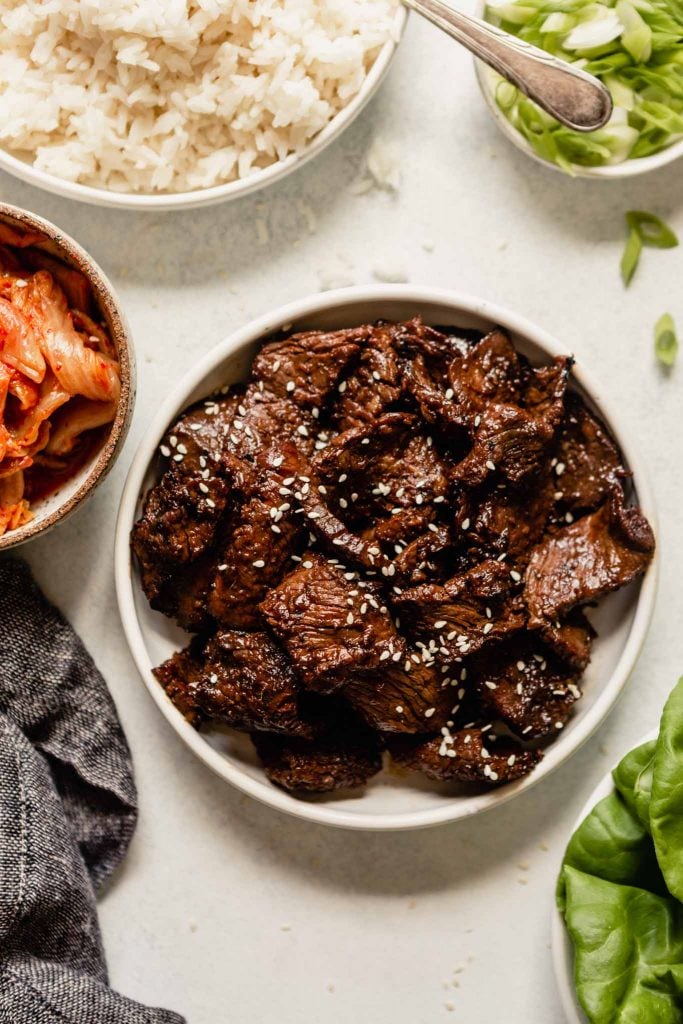 grilled beef in a white bowl set on a table with kimchi, rice, and scallions set around it