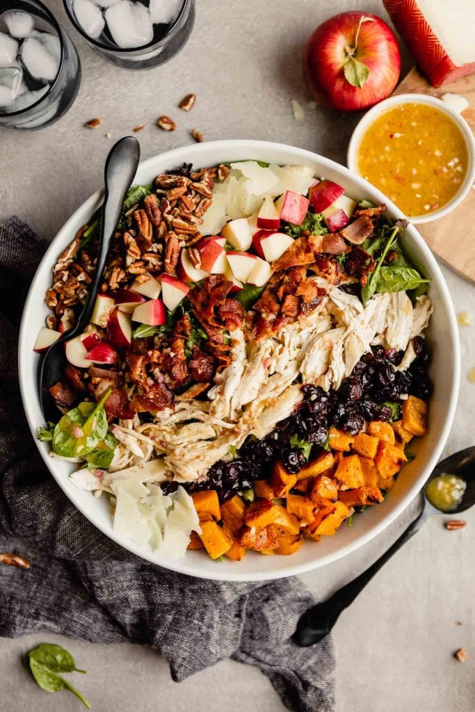 large white bowl filled with dark lettuce greens topped with pecans, apples, Manchego cheese, bacon, chicken, cranberries and squash