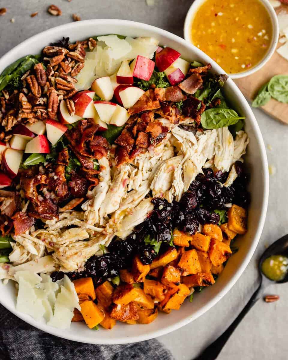 large white bowl filled with dark lettuce greens topped with pecans, apples, Manchego cheese, bacon, chicken, cranberries and squash