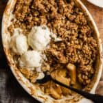 apple crisp in a white oval baking dish set on a wood table. three scoops of ice cream set on top of the crisp with and a black spoon set in the crisp.