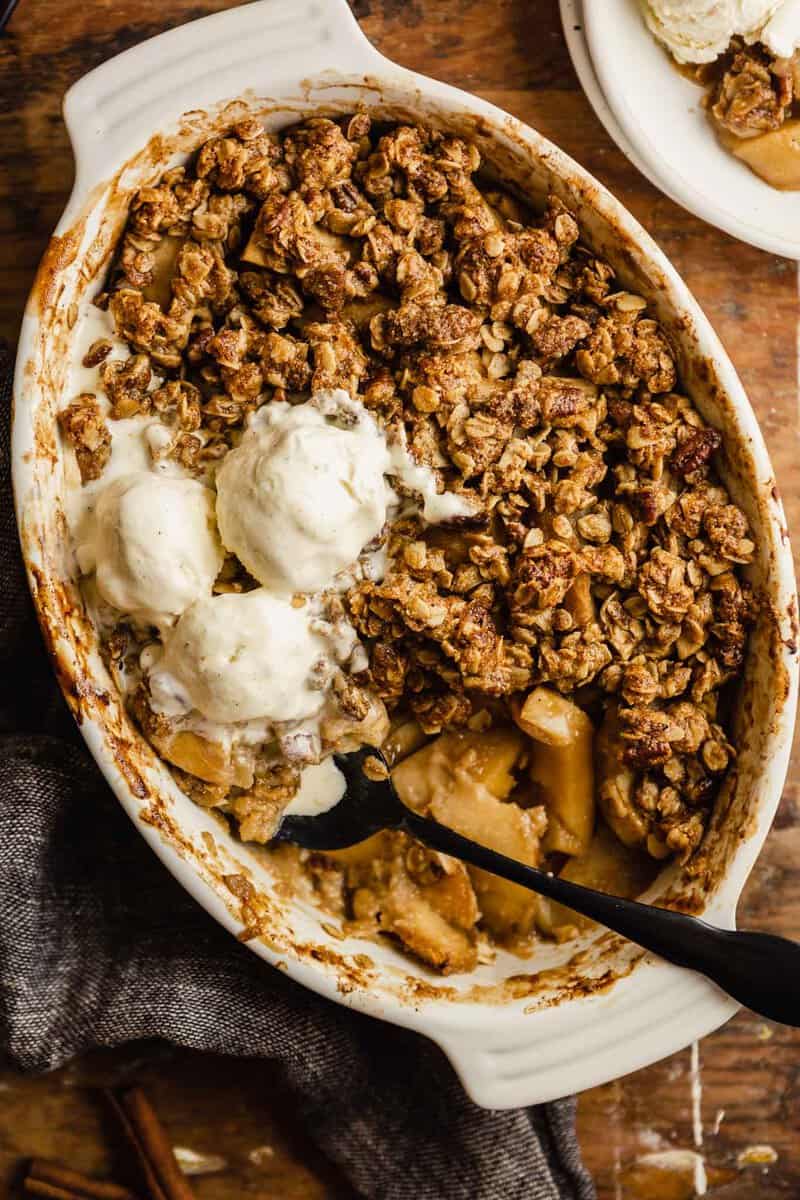 apple crisp in a white oval baking dish set on a wood table. three scoops of ice cream set on top of the crisp with and a black spoon set in the crisp.