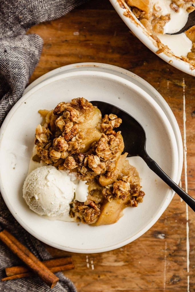 white bowl with melted ice cream and apple crisp set on a wooden table with a black spoon set in bowl