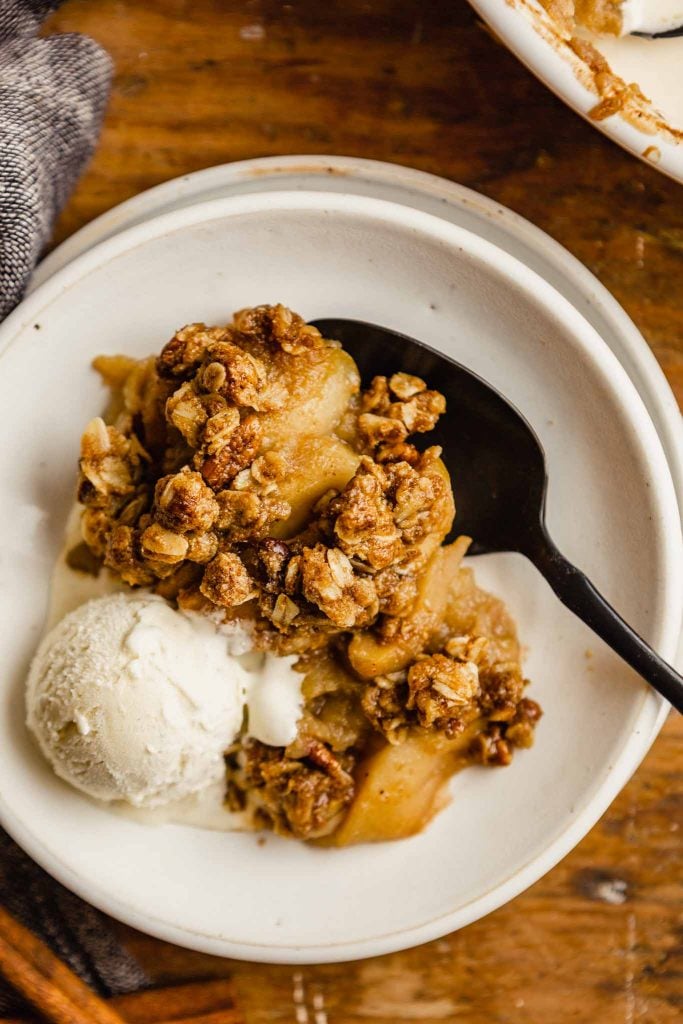 white bowl with melted ice cream and apple crisp set on a wooden table with a black spoon set in bowl