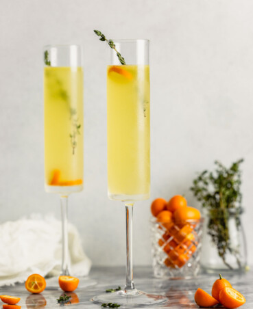 champagne glasses set on a marble table and filled with a yellow drink, kumquats and thyme sprigs