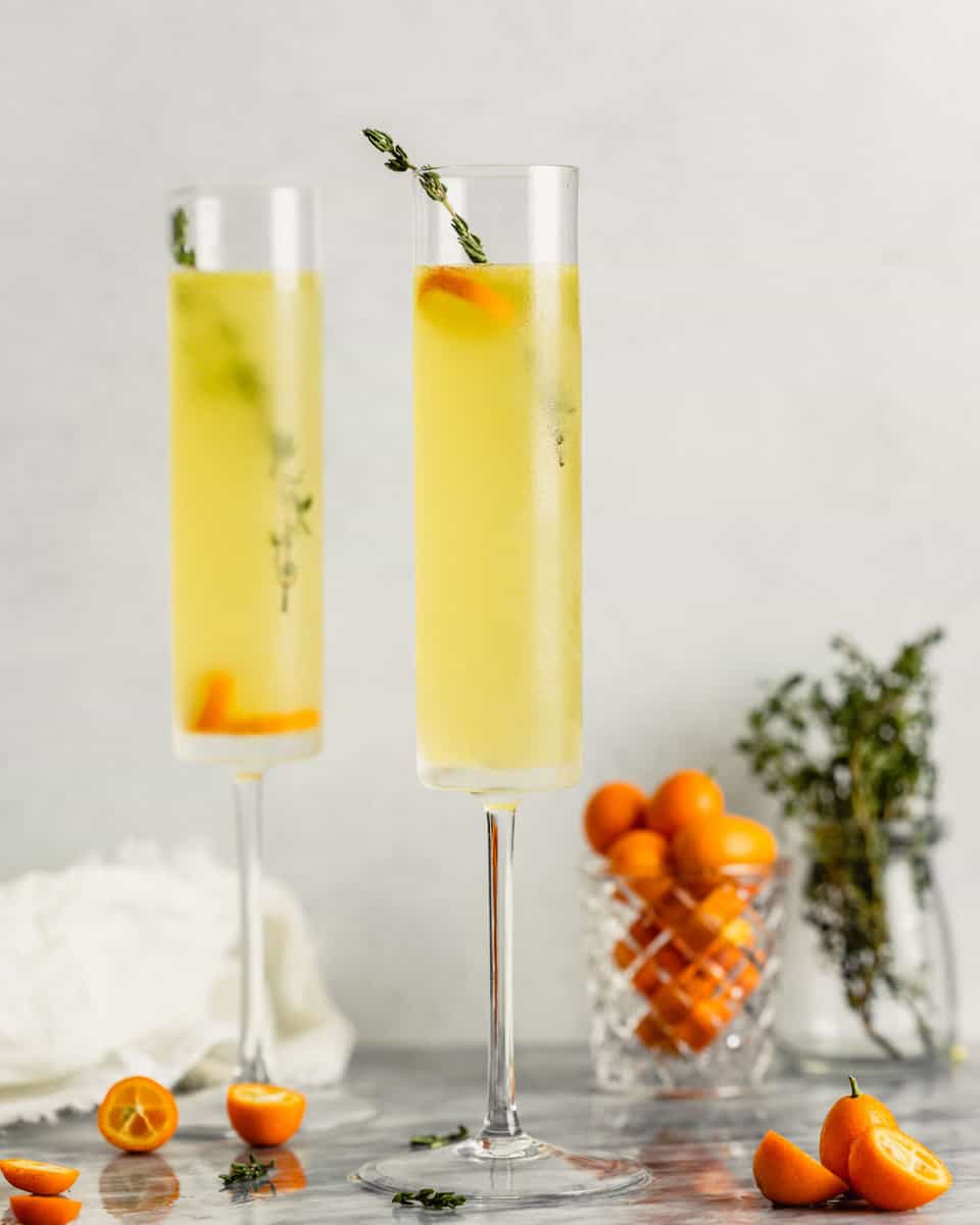 champagne glasses set on a marble table and filled with a yellow drink, kumquats and thyme sprigs