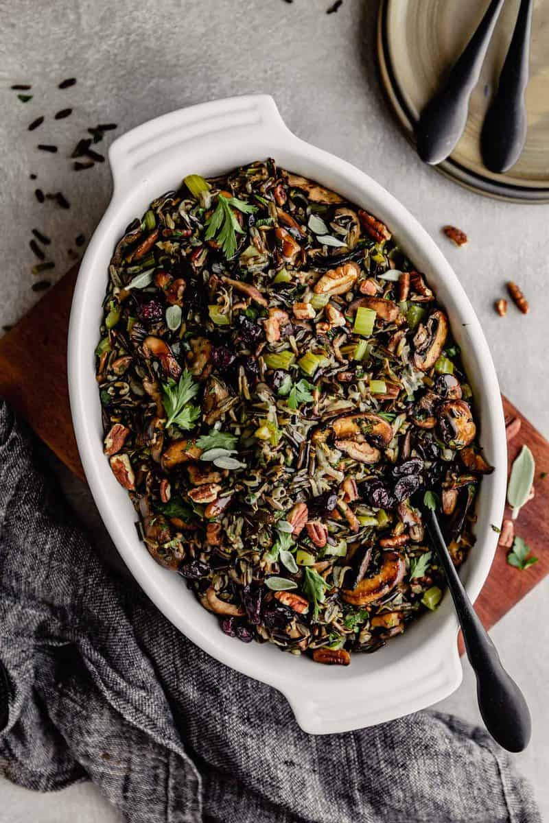 wild rice stuffing in a white oval baking dish set on a wood board on a gray table
