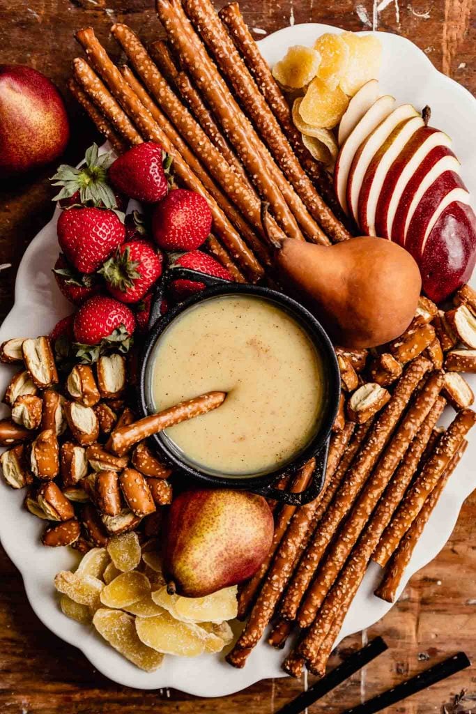 large white platter stacked with pretzels, pears, strawberries, candied ginger with a black pot of fondue in the middle