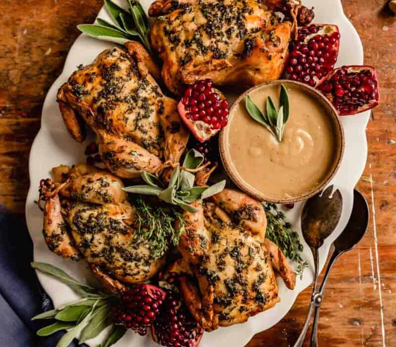 herby-roasted cornish hens on a white platter with herbs and pomegranate pieces