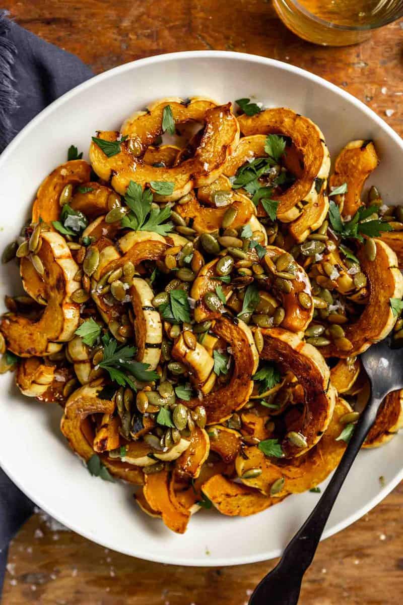 roasted delicata squash slices in a white bowl topped with pepitas, parsley and flaky sea salt.