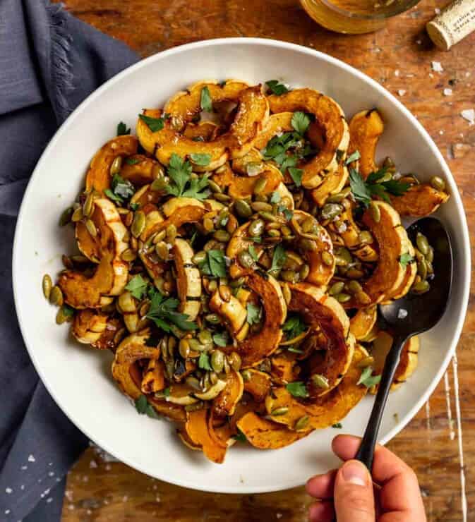 roasted delicata squash slices in a white bowl topped with pepitas, parsley and flaky sea salt.