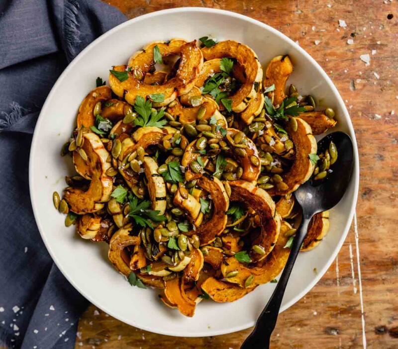 roasted delicata squash slices in a white bowl topped with pepitas, parsley and flaky sea salt.