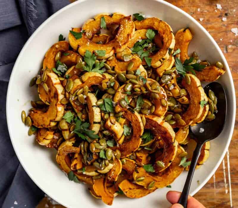 roasted delicata squash slices in a white bowl topped with pepitas, parsley and flaky sea salt.