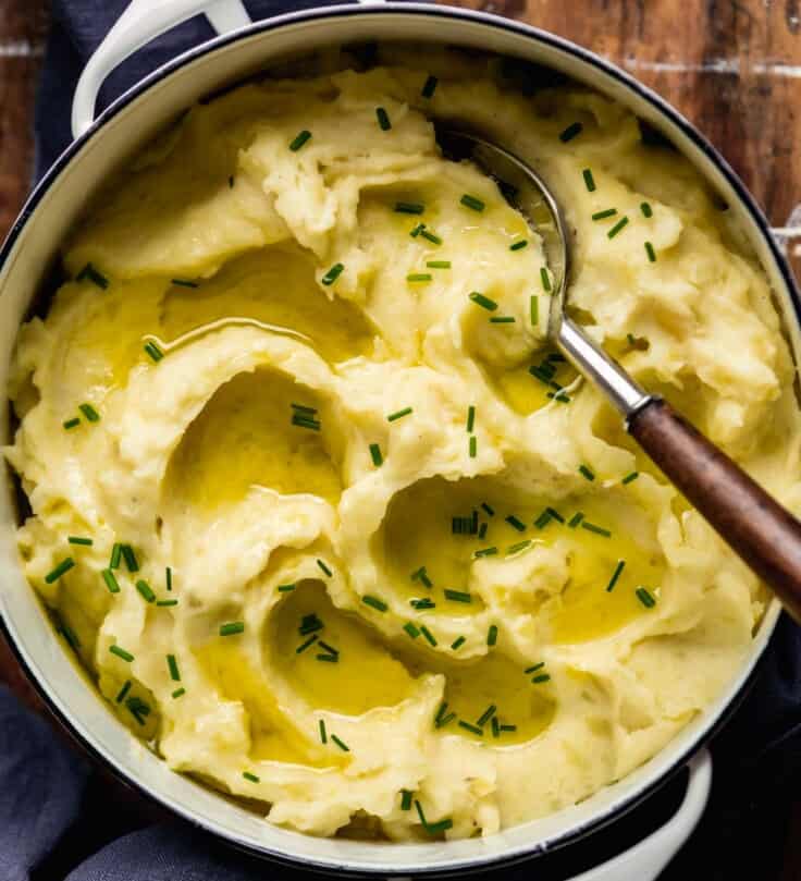 Fluffy mashed potatoes in a white pot set on a wooden table. Olive oil pools on top of potatoes.
