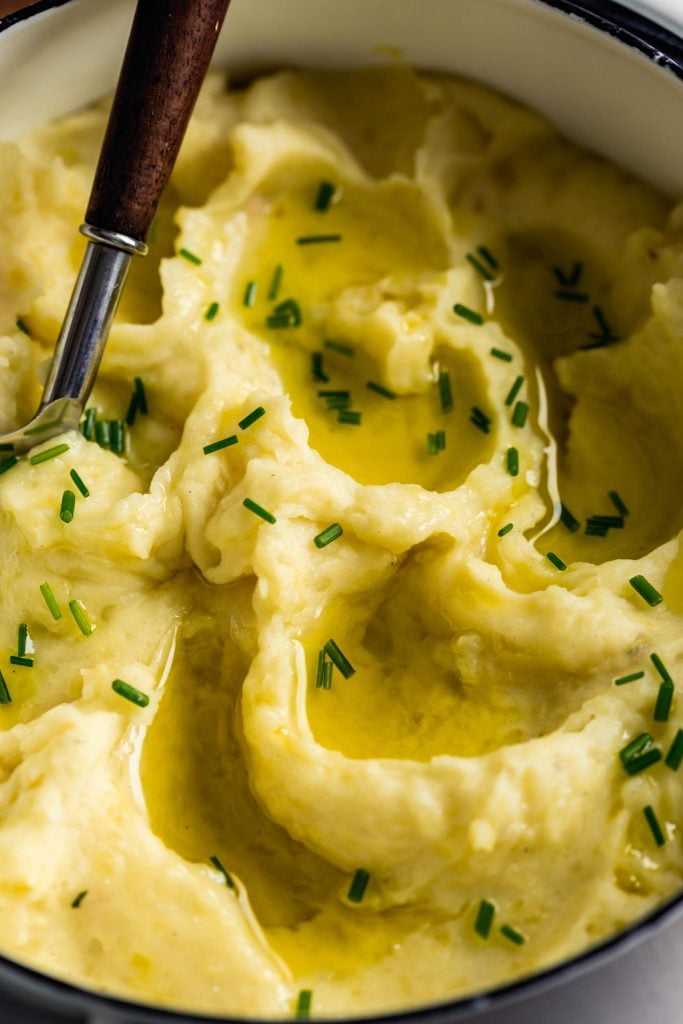 Fluffy mashed potatoes in a white pot set on a wooden table. Olive oil pools on top of potatoes.