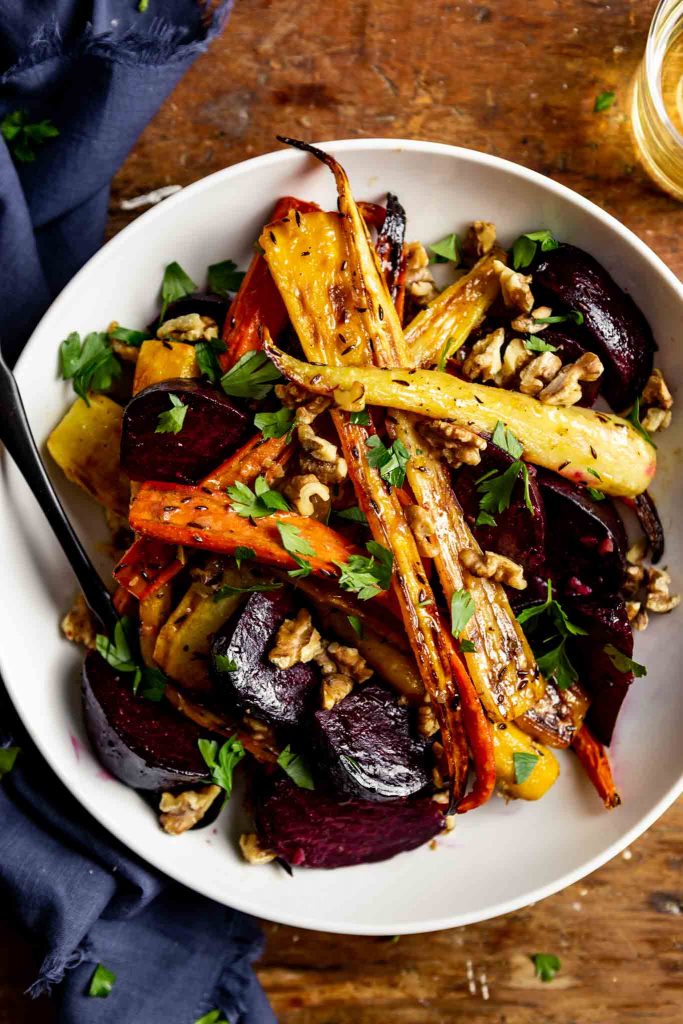 roasted carrots and beets in a white bowl set on a wooden table with a blue napkin and a glass of white wine