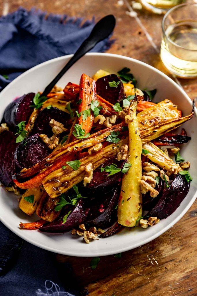 roasted carrots and beets in a white bowl set on a wooden table with a blue napkin and a glass of white wine