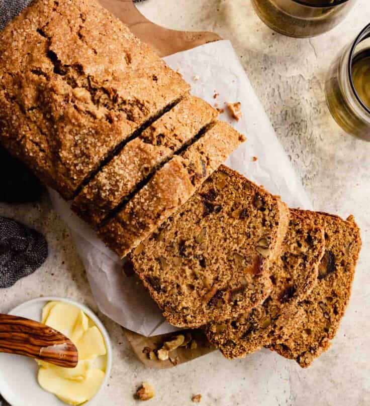 brown bread studded with chopped dried fruit and nuts, set on a parchment-lined wood board set on a table with a butter dish and brown cups set around it