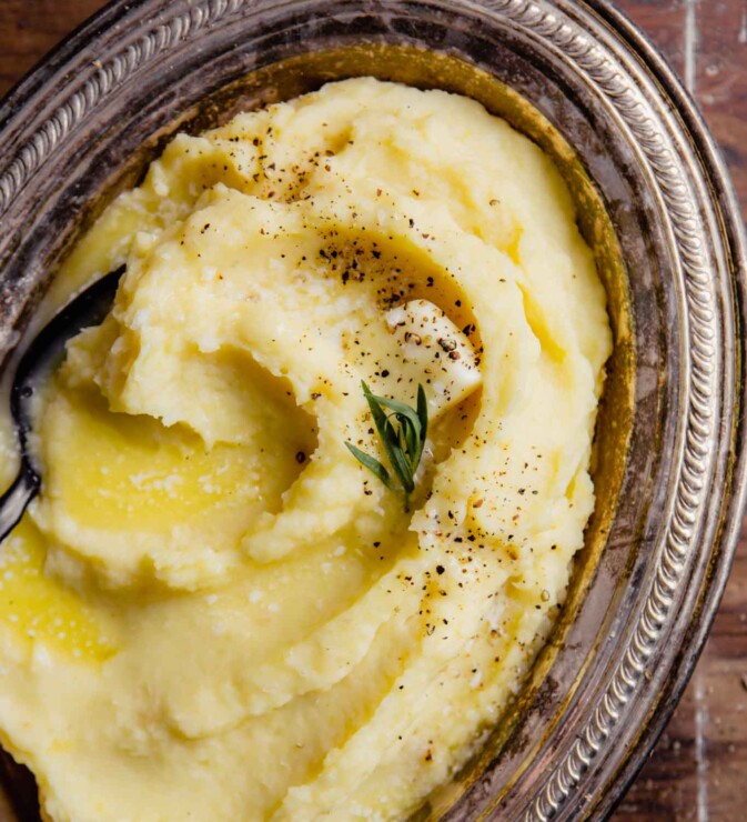 creamy parsnip and potato mash in a silver oval dish with a pad of butter, pepper, salt and tarragon sprinkled over top