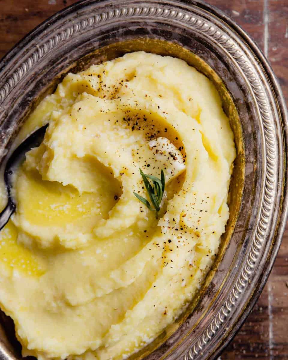 creamy parsnip and potato mash in a silver oval dish with a pad of butter, pepper, salt and tarragon sprinkled over top