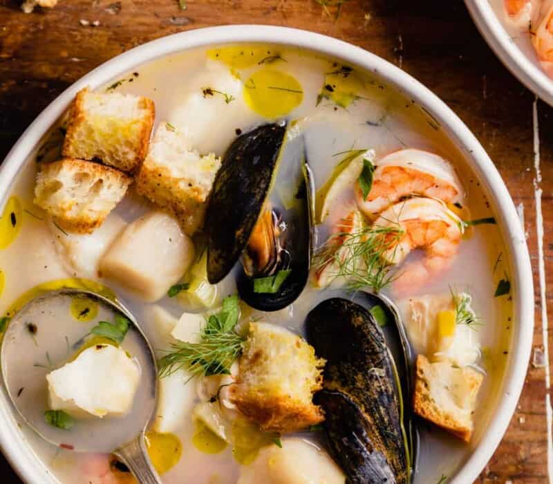 mussels, shrimp, scallops and chunks of white fish in broth with herbs and croutons in a white bowl set on a wood table